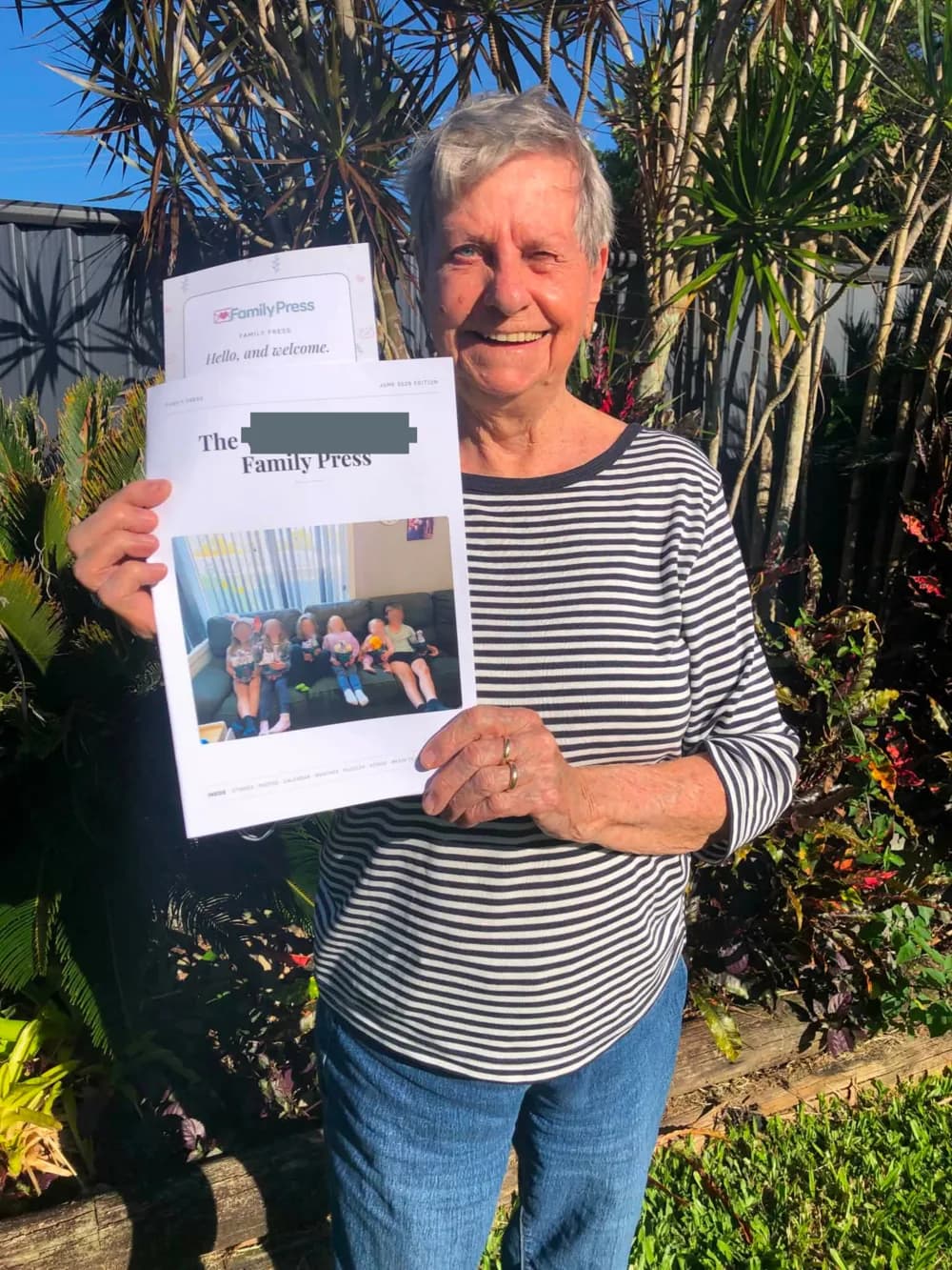Kirsty’s Oma in her sunny garden, smiling as she holds the Family Press newsletter Kirsty made for her